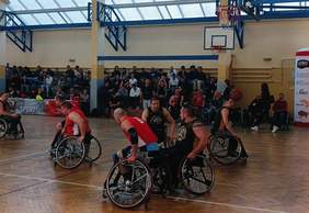 Pokaż zdjęcie: Zdjęcie przedstawia dynamiczną akcję podczas meczu koszykówki na wózkach. Zawodnicy w czerwonych koszulkach i czarnych z żółtymi elementami walczą o piłkę na boisku. Jeden z zawodników wykonuje podanie, a inni gracze są w ruchu. W tle widoczna jest pełna trybuna kibiców oraz banery promocyjne związane z wydarzeniem sportowym.