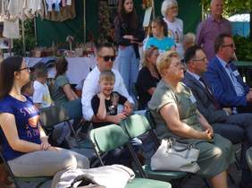 Pokaż zdjęcie: osoby siedzą na wyznaczonych miejscach dla widzów z dala stoiska z rękodziełem wykonanym przez osoby niepełnosprawne