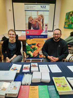 Pokaż zdjęcie: W sali siedzi kobieta i mężczyzna. Uśmiechają się szeroko. Przed nimi ustawiony jest stół, na którym leżą różne informatory poradniki, ulotki, ołówki, długopisy i materiały reklamowe. 