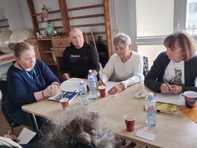 Pokaż zdjęcie: Przy stole siedzi jeden mężczyzna oraz kobiety. Trwają zajęcia informacyjno-doradcze dla osób z niepełnosprawnością. Na stole leżą poradniki, notatki, kubki i woda mineralna. 