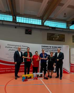 Pokaż zdjęcie: W sali stoi sześć osób: jedna kobieta i pięciu mężyczyzn. Trzech panów trzyma w dłoniach zwycięskie puchary. Na podłodze leżą nagrody, które są przewiązane czerwonymi wstążkami. 