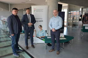 Pokaż zdjęcie: Na zdjęciu stoi 3 mężczyzn. Znajdują się na tle grafiki reklamującej PFRON. Obok nich stoi kanapa, na której rozłożone są ulotki oraz książki