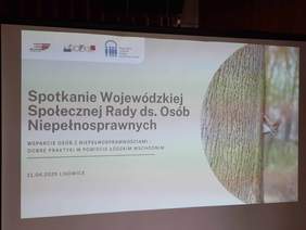 Pokaż zdjęcie: Na tablicy multimedialnej wyświetlony jest tekst: Spotkanie Wojewódzkiej Społecznej Rady ds. Osób Niepełnosprawnych. Wsparcie osób z niepełnosprawnościami – dobre praktyki w Powiecie Łódzkim Wschodnim. 