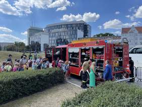 Pokaż zdjęcie: Na placu przy żywopłocie stoi duży pojazd strażacki. Przy nim tłum dzieci. Strażak prezentuje co się znajduję wewnątrz samochodu.