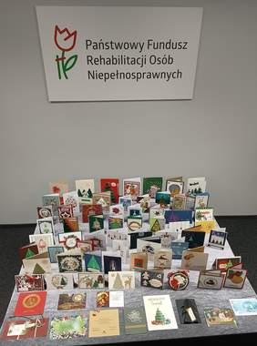 Pokaż zdjęcie: Kilkadziesiąt kartek bożonarodzeniowych ułożonych na stole przykrytym szarym płótnem. W tle widoczny jest przymocowany do ściany duży logotyp Państwowego Funduszu Rehabilitacji Osób Niepełnosprawnych.