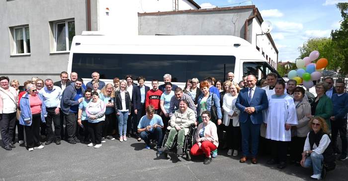 Pokaż zdjęcie: Grupa blisko czterdziestu osób stoi przed białym busem przyozdobionym różnokolorowymi balonami. Wśród zgromadzonych osób znajduje się również ksiądz. W tle biały budynek z płaskim dachem.