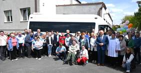 Pokaż zdjęcie: Grupa blisko czterdziestu osób stoi przed białym busem przyozdobionym różnokolorowymi balonami. Wśród zgromadzonych osób znajduje się również ksiądz. W tle biały budynek z płaskim dachem.