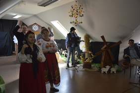 Pokaż zdjęcie: Na scenie stoją artyści, którzy przygotowali świąteczne przedstawienie. Śpiewają i grają na instrumentach. Są przepięknie ubrani. W tle widać dekoracje sceniczne. 