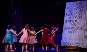 Pokaż zdjęcie: Na scenie grupa dziewcząt ubranych w kolorowe sukienki, tańczy w kółku trzymając się za ręce, z boku widać plakat z informacją o 18 biłgorajskim przeglądzie muzyczno teatralnym