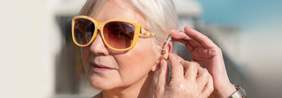 Pokaż zdjęcie: Starsza kobieta zakłada sobie aparat słuchowy na lewe ucho. Ma siwe włosy i żółte przeciwsłoneczne okulary. 