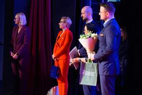 Pokaż zdjęcie: Zdjęcie przedstawia cztery osoby stojące na scenie. Od lewej: kobieta w bordowym garniturze, kobieta w jasnopomarańczowym lub czerwonym garniturze trzymająca niebieską torbę, mężczyzna z brodą i łysą głową, oraz mężczyzna w granatowym garniturze trzymający bukiet kwiatów i zieloną torbę z logo. Za nimi po prawej stronie widać częściowo jeszcze jedną kobietę. Osoby te wydają się być gośćmi lub wręczającymi nagrody. Ubrani są w stroje formalne. Tło jest ciemne, z bordową zasłoną pośrodku.