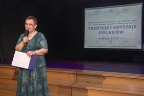 Pokaż zdjęcie: po lewej stronie sceny stoi kobieta w ręku trzyma mikrofon za nią wyświetlony slajd z nazwą konkursu plastycznego. 