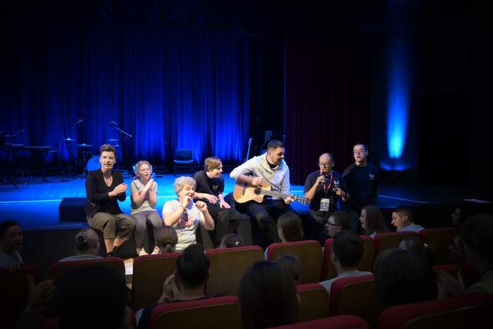 Pokaż zdjęcie: Grupa teatralno-wokalna siedzi na brzegu sceny, blisko publiczności, jedna z osób gra na gitarze. Wszyscy śpiewają.