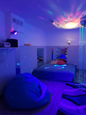 Pokaż zdjęcie: Pokój z kolorowym światłem na suficie tworzącym kolorową rozetę. Na ścianach odbijają się białe punkty. W lewym rogu stoi kanapa przykryta kocem, na niej wyświetla się wąż, po lewej stronie łóżka jest lustro, tak samo u wezgłowia. Na ł