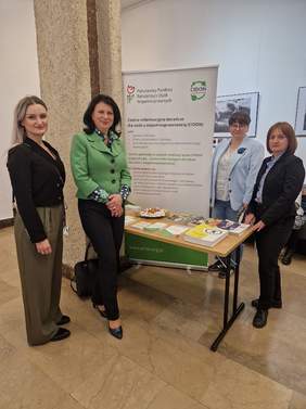 Pokaż zdjęcie: Cztery kobiety stoją obok stoiska informacyjnego. Na stoliku są ulotki i informatory.