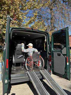 Pokaż zdjęcie: Na parkingu zaparkowany jest samochód przystosowany do przewozu osób z niepełnosprawnością. Tylne drzwi pojazdu są otwarte i we wnętrzu znajduje się pan siedzący na wózku inwalidzkim. W tle widać drzewa i błękitne niebo. 
