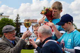 Pokaż zdjęcie: Na podium stoi trzech chłopów. Na same 1 miejscu zawodnik ze spastycznością rąk trzyma kwiaty. Starszy mężczyzna w kaszkiecie i marynarce wręcza mu złoty puchar.