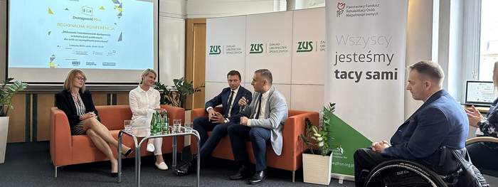 Pokaż zdjęcie: Dwaj mężczyźni w garniturach i dwie kobiety siedzą na dwuosobowych sofach, przed nimi szklany stolik na  którym leża butelki i szklanki, za nimi ekran z prezentacją z logo dostępności. Mężczyzna w jasnej marynarce trzyma mikrofon. Obok niego st