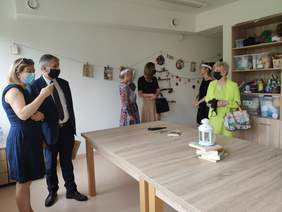 Pokaż zdjęcie: Pomieszczenie pracowni rękodzieła w środku znajduje się 6 osób w tym jeden mężczyzna rozmawiają i rozglądają się na środku stoi duży drewniany stół przy jednej ścianie stoi szafa z regałami na których ustawione są różne materiały do 