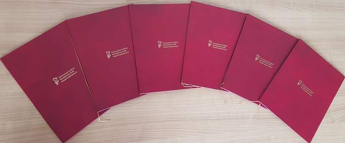 Pokaż zdjęcie: Sześć teczek w kolorze bordowym z logo Państwowego Funduszu Rehabilitacji Osób Niepełnosprawnych jest ułożonych na stole. Teczki zostały położone w taki sposób, że zachodzą jedna na drugą.