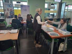 Pokaż zdjęcie: W sali obsługi klienta widoczne są stoiska Państwowego Funduszu Rehabilitacji Osób Niepełnosprawnych oraz Narodowego Funduszu Zdrowia. Przy stoisku Oddziału Wielkopolskiego PFRON stoi kobieta, której pracownik Oddziału udziela informacji. Na stoliku wyłożone są ulotki i publikacje na temat programów i zadań realizowanych przez PFRON.
