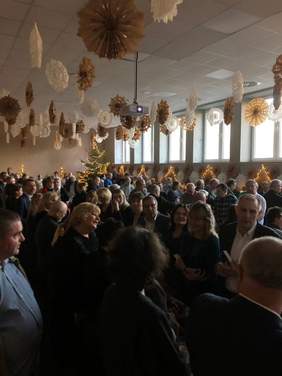 Pokaż zdjęcie: Przy stołach stoją ludzie. Uśmiechają się do siebie. Wszyscy trzymają w dłoniach opłatki. 