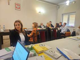 Pokaż zdjęcie: Na pierwszym planie pracownik Oddziału Świętokrzyskiego PFRON siedzący za stołem, przed nim laptop z wyświetlaną prezentacją; w tle uczestnicy spotkania