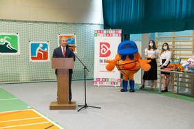 Pokaż zdjęcie: fragment sali gimnastycznej przy mikrofonie stoi mężczyzna w garniturze za nim stoi nowa maskotka szkoły pomarańczowa piłka w granatowej czapce i butach