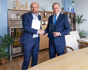 Dwaj mężczyźni, w garniturach, stoją, uśmiechają się, ściskają sobie dłonie, przekauzją sobie dokumenty, patrzą w obiektyw