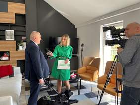 Pokaż zdjęcie: Mężczyzna w ciemnym garniturze trzymający uprząż z psem asystującym rozmawia z piękną kobietą w blond włosach, w zielonej, luźnej sukience. Obok stoi ubrany w szarą bluzę i jeansy, łysy mężczyzna patrzący przez kamerę stojącą na statyw