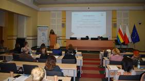 Pokaż zdjęcie: Na zdjęciu sala konferencyjna z uczestnikami siedzącymi przy stołach. Przy mównicy stoi Elżbieta Pomian, pracownik PFRON, wygłaszająca prelekcję. Za nią na ekranie wyświetlana jest prezentacja z tytułem "Cel programu", dotyczącym wyrównywania szans osób z niepełnosprawnościami. Na podium za stołem prezydialnym siedzą dwie kobiety przy laptopach. W tle znajdują się trzy flagi: Polski, województwa podlaskiego i Unii Europejskiej. Widoczna jest także tablica z logo Regionalnego Ośrodka Polityki Społecznej w Białymstoku.