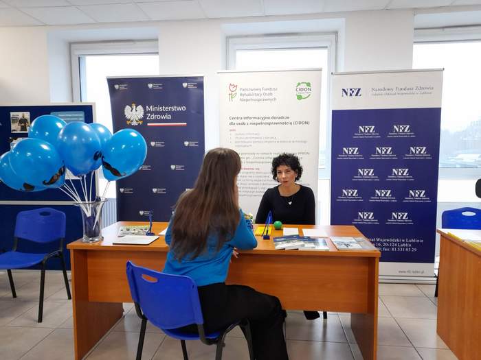 Pokaż zdjęcie: Przy biurku siedzą dwie kobiety, jedna z nich plecami do aparatu. W tle banery z logo PFRON i CIDON. 