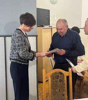 Pokaż zdjęcie: Kobieta w czarno-białej, wzorzystej marynarce wręcza dyplom oraz nagrodę uśmiechniętemu mężczyźnie w ciemnej koszuli. Jest to moment nagrodzenia laureata Konkursu Plastycznego „Sztuka Osób Niepełnosprawnych”. Na pierwszym planie po prawej widać oparcie zdobionego, drewnianego krzesła, a w tle jasną ścianę z szafką i telewizorem.