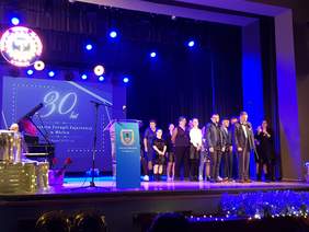 Ciemno niebieskie tło. Na scenie po prawej stronie uczestnicy śpiewają, z lewej mężczyzna gra na pianinie