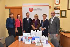 Pokaż zdjęcie: Uczestnicy inauguracyjnego spotkania w Starostwie Powiatowym w Limanowej. Kobiety i mężczyźni stoją na tle rollupu z napisem Powiat Limanowski. Przed nimi na stole przykładowa zawartość paczki: akcesoria ochrony osobistej, środki dezynfekcyjne, pr