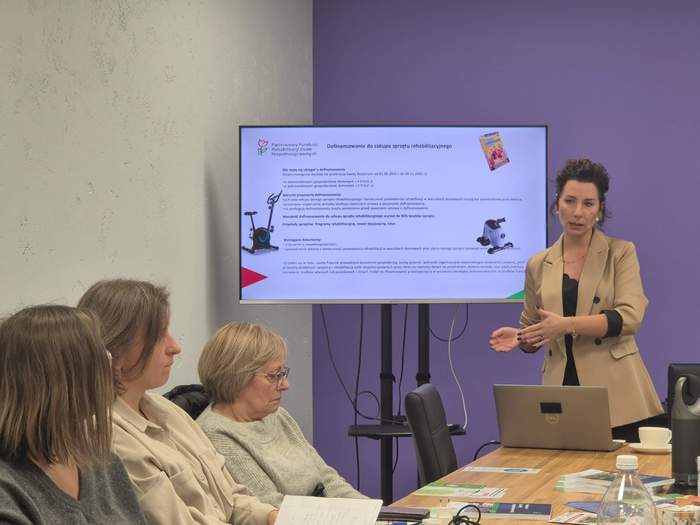 Pokaż zdjęcie: Kobieta w beżowej marynarce stoi przy dużym monitorze i wygłasza prelekcję do osób siedzących przy stole.