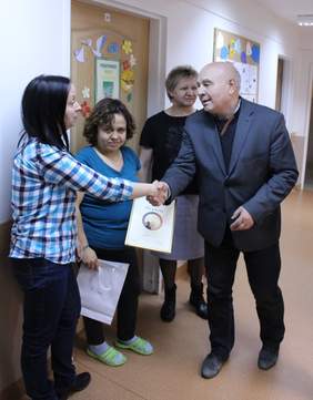 Pokaż zdjęcie: W budynku stowarzyszenia zdjęcie czterech osób pozujących na korytarzu. Mężczyzna ściska dłoń kobiecie i przekazuje gratulacje, za nimi stoi kobieta nagrodzona z dyplomem oraz starsza kobieta prezes stowarzyszenia.