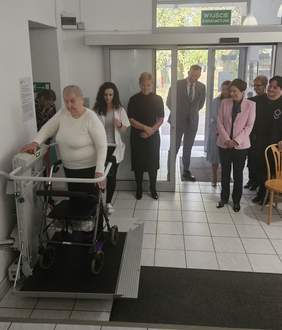 Pokaż zdjęcie: Grupa ośmiu osób znajdująca się na parterze budynku przygląda się kobiecie z balkonikiem, która testuje nową platformę przyschodową. W tle otwarte drzwi prowadzące na zewnątrz budynku. Nad nimi widnieje napis: „Wyjście ewakuacyjne”.