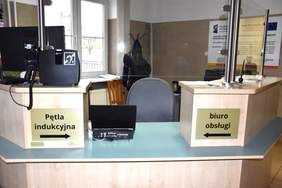 Pokaż zdjęcie: Lada z tabliczkami: „biuro obsługi” i „pętla indukcyjna”. Za ladą są dwa krzesła, monitor, grzejnik, wieszak z kurtką oraz okno. Na ścianie zawieszona jest tablica informująca o dofinansowaniu rozwoju systemów informatycznych ze środków unijnych.