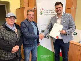 Pokaż zdjęcie: Dwóch mężczyzn podaje sobie dłoń, jeden z nich trzyma w ręku teczkę z logo PFRON. Obok jednego z mężczyzn kobieta w czapce. Za nimi baner reklamujący PFRON oraz duża szafa.