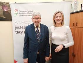 Pokaż zdjęcie: Do zdjęcia pozują dwie osoby: kobieta i mężczyzna. Uśmiechają się. Za nimi stoi baner Państwowego Funduszu Rehabilitacji Osób Niepełnosprawnych. 