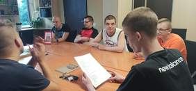 Pokaż zdjęcie: sześciu młodych mężczyzn siedzi przy stole oglądają książki czytają lub słuchają