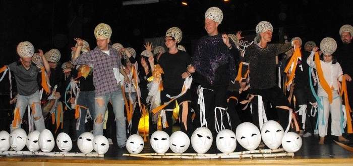 Pokaż zdjęcie: Na scenie stoi kilkanaścioro młodych ludzi. Machają szarfami. Mają założone coś okrągłego i prześwitującego na głowach. Przed nimi na podłodze leżą białe maski.