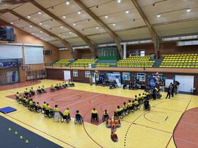 Widok hali sportowej z góry. Osoby poruszające się na wózkach tworzą duży okrąg na środku boiska. W tle znajdują się trybuny, dekoracje z balonów oraz stanowiska rozmieszczone przy ścianie hali.)