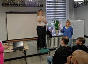 Pokaż zdjęcie: 2.	Kilka osób siedzi w sali wykładowej, trzy kobiety stoją z przodu sali. Kobieta po prawej stronie opowiada o formach wsparcia, kobieta w środku stoi na podwyższeniu i  tłumaczy słowa na język migowy, kobieta po lewej stronie przygląda się. W sali znajduje się stół, na ścianie rzutnik, okno, po prawej stronie tablica. Nad tablicą i rzutnikiem dużo pucharów.