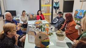 Pokaż zdjęcie: Grupa osób siedzi przy stole, na stole ustawione są kolorowe rzeźby 