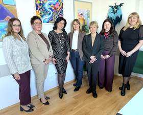 Siedem kobiet pozuje do zdjęcia. Wszystkie są elegancko ubrane i uśmiechnięte. Na ścianie za nimi widać fragmenty obrazów.