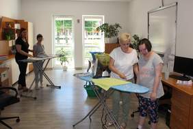 Pokaż zdjęcie: W sali przy deskach do prasowania stoją dwie dziewczynki ,a przy nich dwie kobiety, które uczą je prasowania