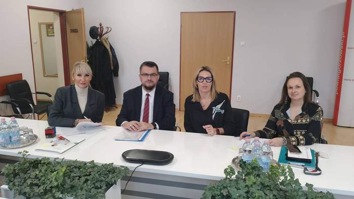 Pokaż zdjęcie: Przy stoliku siedzą osoby podpisujące umowę, są na nim dokumenty, widać kwiaty, otwarte drzwi