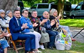 Pokaż zdjęcie: Na ławeczkach w plenerze siedzą zaproszeni goście. Mężczyzna w garniturze postawi przed sobą dwa kosze z cukierkami.